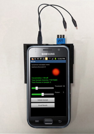 :img Arrangement used for mobile phone based ECL sensing. The audio jack supplies the potential to the paper microfluidic sensor while the resultant emission is detected by the camera in video mode. Both the excitation and detection processes are controlled by a software application which can also transmit the results via e-mail. The black plastic sleeve surrounding the top of the phone holds the sensor adjacent to the camera and blocks ambient light., 60%
